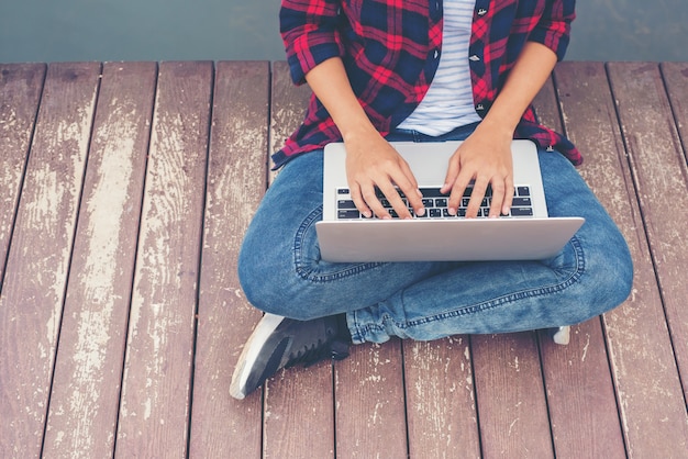 Young hipster hands typing laptop sitting on pier lake, relaxing