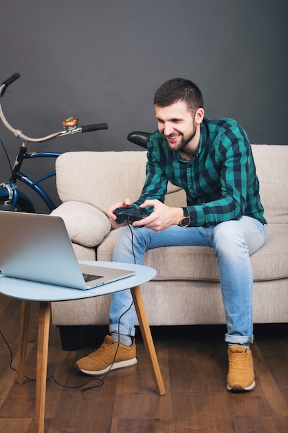 Young hipster handsome bearded man sitting on couch at home, playing video game on notebook