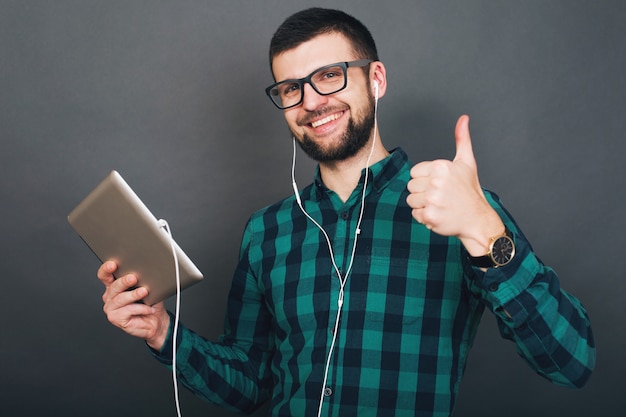 Young hipster handsome man on grey background holding tablet listening to music on earphones talking online happy smiling green checkered shirt eyeglasses, positive mood