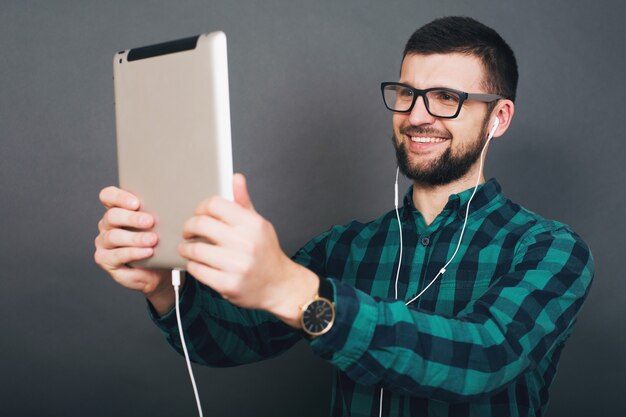 Young hipster handsome man on grey background holding tablet listening to music on earphones talking online