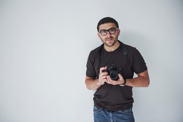 A young hipster man in eyepieces holds a DSLR camera in hands standing against a white wall