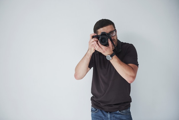 A young hipster man in eyepieces holds a DSLR camera in hands standing against a white wall