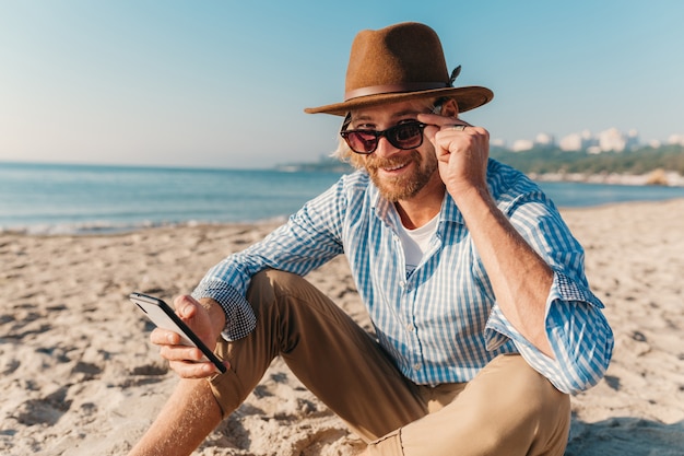Young hipster man sitting on beach by sea on summer vacation, boho style outfit, holding using internet on smartphone