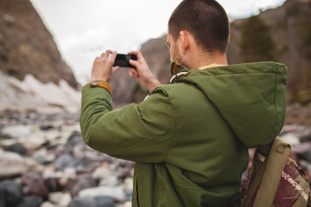 Young hipster man, taking pictures using smartphone, wild nature, winter vacation, hiking