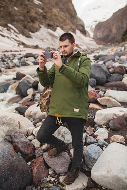 Young hipster man, taking pictures using smartphone, wild nature, winter vacation, hiking