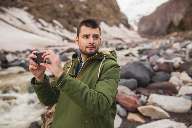 Young hipster man, taking pictures using smartphone, wild nature, winter vacation, hiking