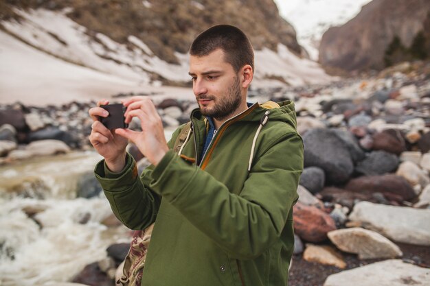 Young hipster man, taking pictures using smartphone, wild nature, winter vacation, hiking