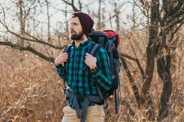 Young hipster man traveling with backpack in autumn forest wearing checkered shirt and hat, active tourist, exploring nature in cold season