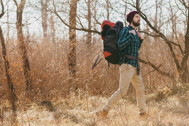 Young hipster man traveling with backpack in autumn forest wearing checkered shirt and hat, active tourist walking, exploring nature in cold season