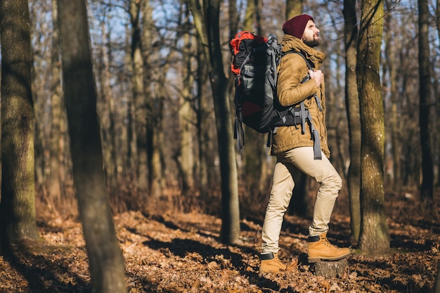 Young hipster man traveling with backpack in autumn forest wearing warm jacket and hat, active tourist, exploring nature in cold season