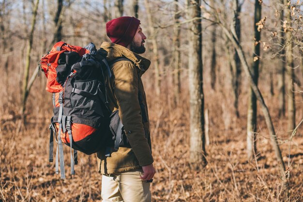 Young hipster man traveling with backpack in autumn forest wearing warm jacket and hat, active tourist, exploring nature in cold season