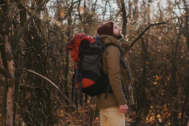Young hipster man traveling with backpack in autumn forest wearing warm jacket and hat, active tourist, exploring nature in cold season