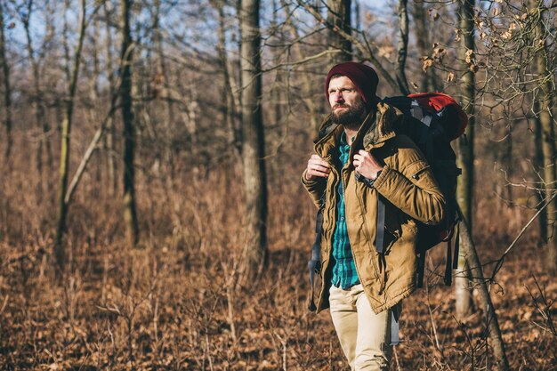 Young hipster man traveling with backpack in autumn forest wearing warm jacket and hat