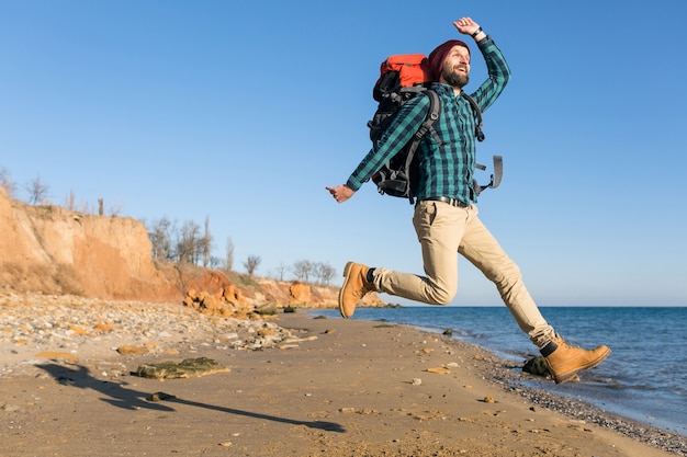 Young hipster man traveling with backpack in autumn sea coast wearing warm jacket and hat