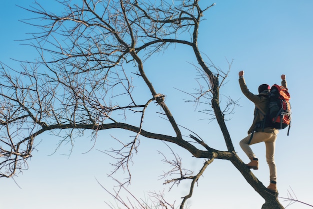 Молодой хипстерский человек путешествует с рюкзаком, стоит на дереве против неба, в теплой куртке, активный турист, исследует природу в холодное время года