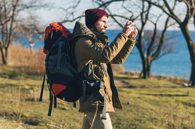 Young hipster man traveling with backpack wearing warm jacket and hat, active tourist, taking pictures on mobile phone, exploring nature in cold season