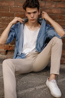 若いヒップスターは彼の青いデニムのボタンアップシャツに座って、レンガの壁の背景にラベルのない白い綿のtシャツを示しています