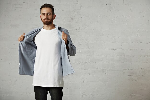 young hipster taking off his faded blue denim button up shirt showing an unlabeled white cotton t-shirt on brick wall