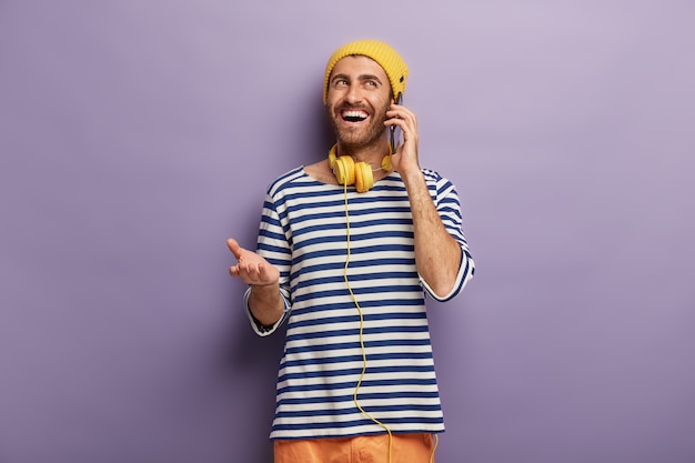 Free photo young hipster talks casually with friend via smartphone, discusses something funny happened with him, has happy face expression, wears stylish outfit, listens music in headphones. communication
