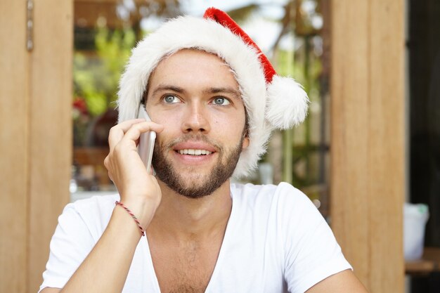 Young hipster with stylish beard wearing red Santa Claus hat holding smart phone