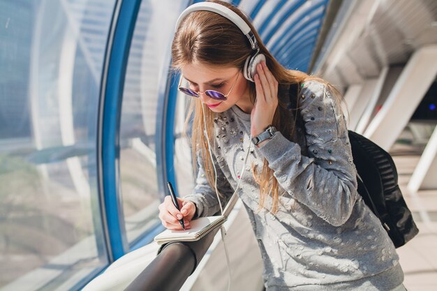 Young hipster woman in casual outfit having fun listening to music in headphones, wearing sweater and sunglasses, student making notes