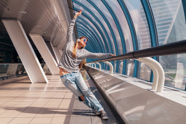 Young hipster woman dancing having fun in urban modern building dressed in casual outfit listening to music in headphones