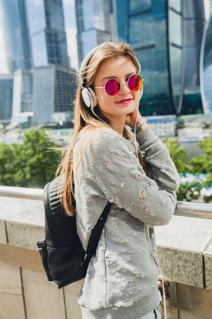 Young hipster woman having fun in street listening to music on headphones, wearing pink sunglasses and backpack, spring summer urban style