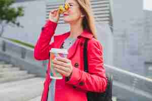 無料写真 ピンクのコートを着た若いヒップスターの女性、バックパックとヘッドフォンで音楽を聴いているコーヒーと通りのジーンズ