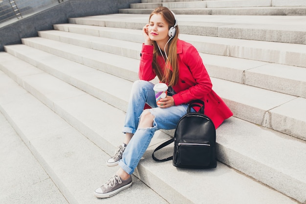 Young hipster woman in pink coat, jeans sitting in street with backpack and coffee listening to music on headphones, big city urban spring style trend