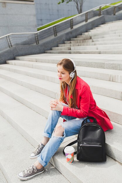 Young hipster woman in pink coat, jeans sitting in street with backpack and coffee listening to music on headphones, big city urban spring style trend