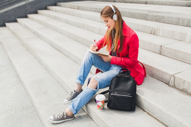 Young hipster woman in pink coat, jeans sitting in street with backpack and coffee listening to music on headphones, student making notes