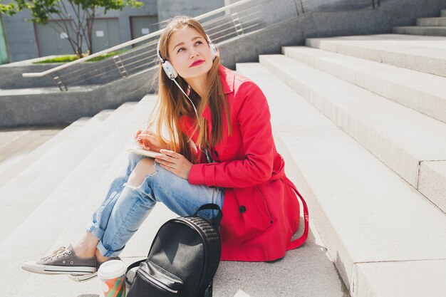 Young hipster woman in pink coat, jeans sitting in street with backpack and coffee listening to music on headphones, student making notes