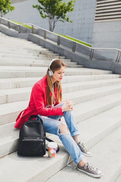 Young hipster woman in pink coat, jeans sitting in street with backpack and coffee listening to music on headphones