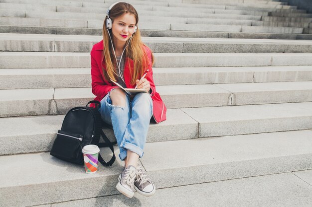 Young hipster woman in pink coat, jeans sitting in street with backpack and coffee listening to music on headphones