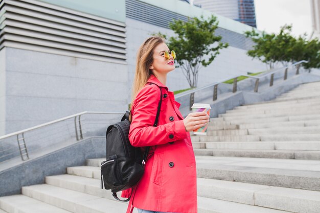 Young hipster woman in pink coat, jeans in street with backpack and coffee listening to music on headphones, wearing sunglasses