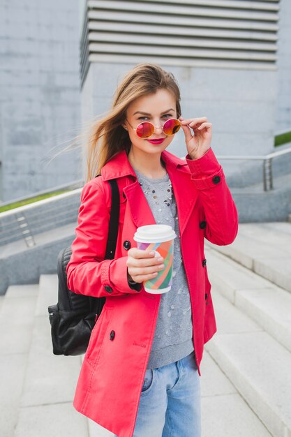 Молодая хипстерская женщина в розовом пальто, джинсах на улице с рюкзаком и кофе, слушая музыку в наушниках, в темных очках