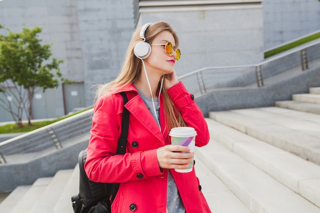 Young hipster woman in pink coat, jeans in street with backpack and coffee listening to music on headphones, wearing sunglasses