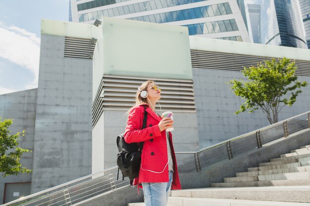 핑크 코트에 젊은 힙 스터 여자, 선글라스를 착용하고 헤드폰에서 음악을 듣고 배낭과 커피 거리에서 청바지