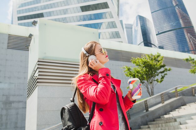 Young hipster woman in pink coat, jeans in street with backpack and coffee listening to music on headphones, wearing sunglasses