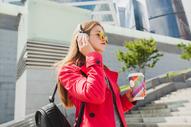 サングラスをかけているピンクのコート、ジーンズのバックパックとコーヒーをヘッドフォンで音楽を聞いて通りの若い流行に敏感な女性