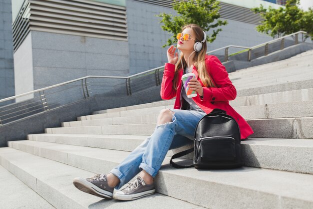 Young hipster woman in pink coat, jeans in street with backpack and coffee listening to music on headphones, wearing sunglasses