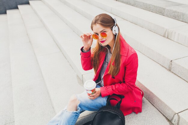 Young hipster woman in pink coat, jeans in street with backpack and coffee listening to music on headphones, wearing sunglasses