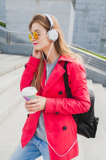 Young hipster woman in pink coat, jeans in street with backpack and coffee listening to music on headphones