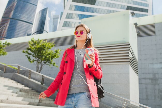 Молодая хипстерская женщина в розовом пальто, джинсах на улице с рюкзаком и кофе, слушает музыку в наушниках