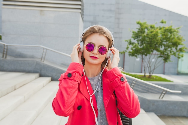 ピンクのコートを着た若いヒップスターの女性、ヘッドフォンで音楽を聴いてコーヒーとストリートのジーンズ