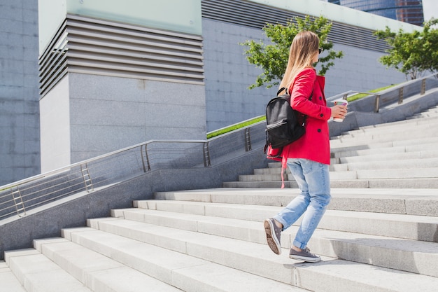 Young hipster woman in pink coat, jeans walking on stairs in street with backpack and coffee listening to music on headphones, wearing sunglasses