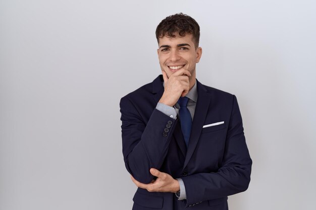 Молодой латиноамериканский деловой человек в костюме и галстуке уверенно смотрит в камеру, улыбаясь со скрещенными руками и поднятой рукой на подбородке, думая позитивно