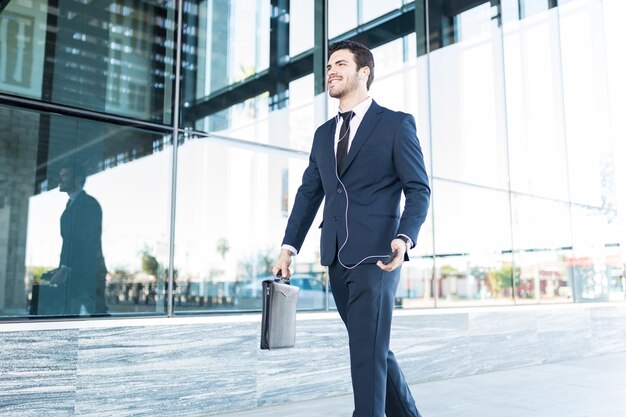 Young Hispanic businessman holding briefcase while enjoying favorite music via earphones in city