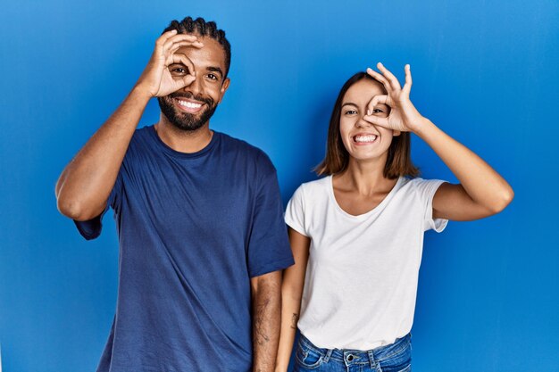 Young hispanic couple standing together doing ok gesture with hand smiling, eye looking through fingers with happy face.