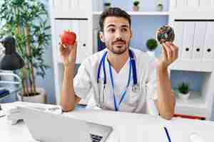 Free photo young hispanic dietitian man holding doughnut and apple smiling looking to the side and staring away thinking.